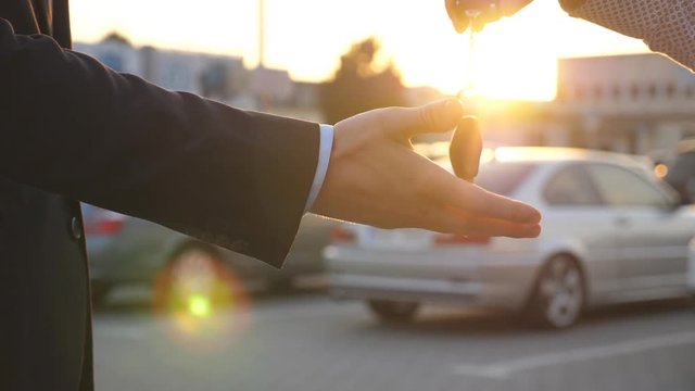 Male hands giving keys of car to his friend with sun flare at background. Arm of businessman passed car key. Handshake between two business men outdoor. Close up Slow motion