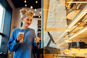 Thoughtful blond woman in blue sweater with latte coffee in hand choosing a cheesecake while standing in front showcase in a coffee or bakery shop.