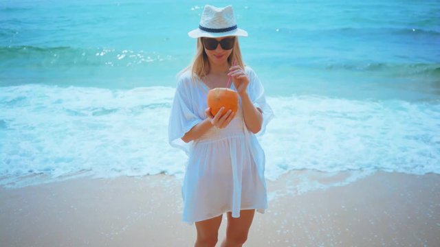 Woman drinks coconut water and walking on the ocean beach under blue sky in sunny day. Seascape view with palms.