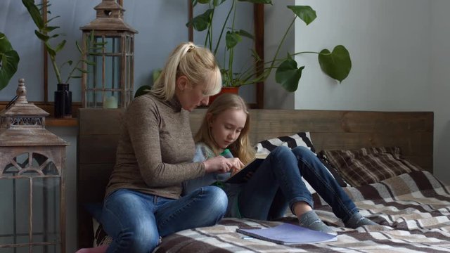 Little school girl learning to read with help of attentive mother while sitting on bed at home. Caring mom teaching her cute daughter to read and correcting spelling mistake while reading a book.