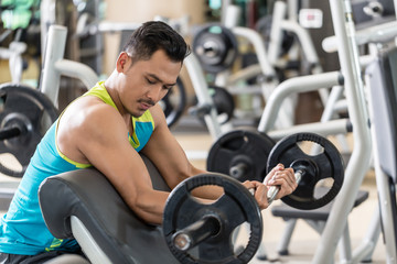 Young handsome man exercising bicep curls with the E-Z barbell while sitting down at preacher curl bench during upper-body workout routine