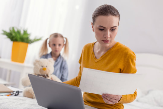 Only work. Attractive focused ambitious mother looking through document while posing on the blurred background and paying no attention to daughter