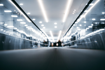 Shooting with very shallow depth of field of moving walkway in bright airport terminal interior; travelator trough contemporary departure area of railway station depot