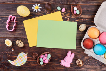 Easter composition and blank cards. Easter eggs, sweets, paper cutouts with yellow and green empty card on brown wooden background, top view.