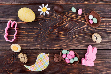 Easter handmade decor on wooden background. Easter festive frame from chocolate, eggs, candies, rabbit molds, papercut bird on brown wooden background, top view. Easter festive mood.