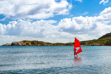 Fototapeta premium Windsurf with red sail rides on sea on background rocky shore. View from sunny Livadi beach in resort village Bali. Rethymno, Crete Greece. Extreme water sports as active recreation on Sunny vacations