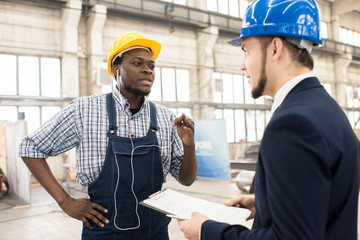 Bearded African American factory worker wearing hardhat and overall discussing results of accomplished work with his superior while standing at production department