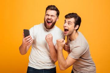 Image of two bachelors screaming and rejoicing win of football team while watching game on mobile phone, isolated over yellow background - obrazy, fototapety, plakaty