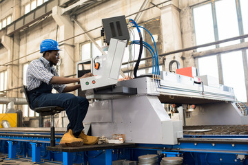 Profile view of young African American worker wearing overall and checked shirt operating machine unit at production department of modern plant