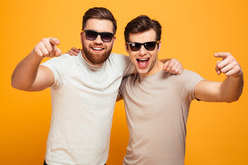 Two happy men in casual t-shirts and sunglasses hugging and pointing fingers on camera meaning hey you, isolated over yellow background
