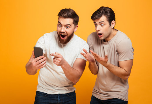 Image of two bachelors screaming and gesturing while watching game on mobile phone, isolated over yellow background