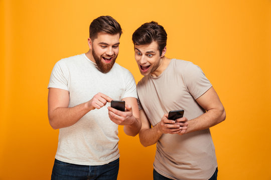 Two joyful men in casual talking and looking at mobile phones in their hands, isolated over yellow background