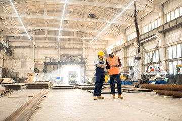 Naklejka na ściany i meble African American engineer wearing reflective vest and hardhat using digital tablet while discussing joint work with his colleague, interior of spacious production department of plant on background