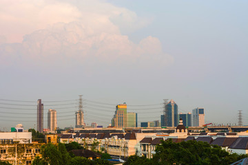 Fototapeta premium A cityscape with tall buildings, power lines, and trees in the foreground. The sky is a pale blue with some white clouds.