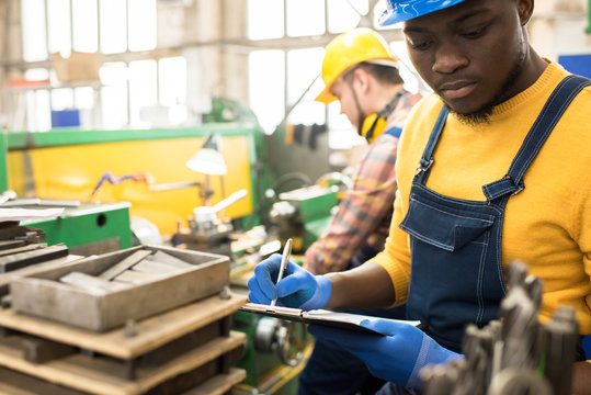 African American inspector wearing hardhat and overall taking notes while carrying out quality control at spacious production department of modern plant, silhouette of lathe operator behind him