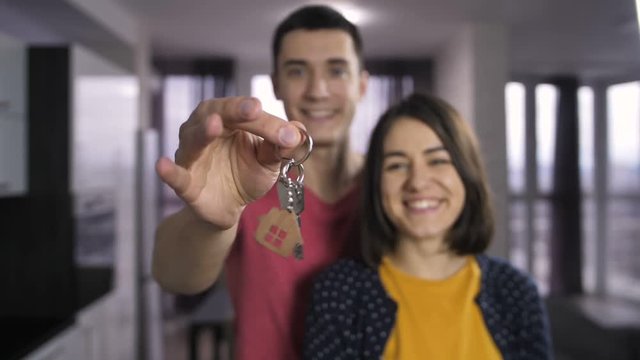 Portrait of happy married couple standing indoors and shaking keys to new house. Male hand holding keys and offering it to the camera. Slow motion. Keys with wooden house shaped key ring.