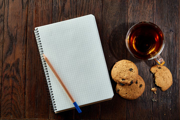 Cup of tea with cookies, workbook and a pencil on a wooden background, top view