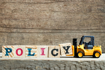 Yellow toy forklift hold letter block Y to complete word policy on wood background