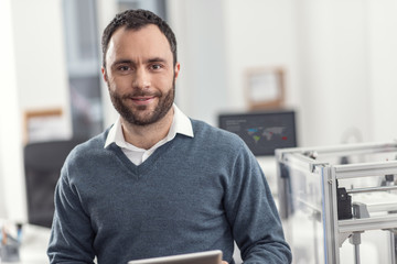 Upbeat worker. The portrait of a joyful young engineer standing near a 3D printer in his office, holding a tablet and posing for the camera, smiling pleasantly
