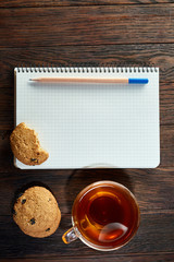 Cup of tea with cookies, workbook and a pencil on a wooden background, top view