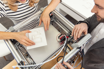 Refinement needed. The top view of pleasant colleagues inserting a half-made model of a house into the 3D printer and being about to proceed with its creation