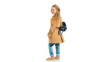 Rear view of smiling child in glasses wearing trench coat and holding bag isolated on white