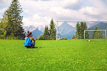 young woman tourist with a backpack and glasses resting on green grass and admiring the alpine mountains and meadows