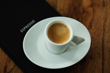 A cup of espresso standing on black book with word espresso on the cover. Wooden Background.