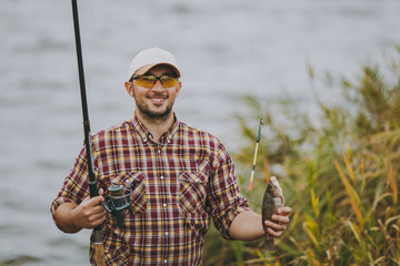 Young unshaven man in checkered shirt, cap and sunglasses pulled out fishing pole with caught fish...