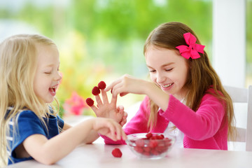 Two pretty little girls eating raspberries at home. Cute children enjoying their healthy fresh organic fruits and berries.