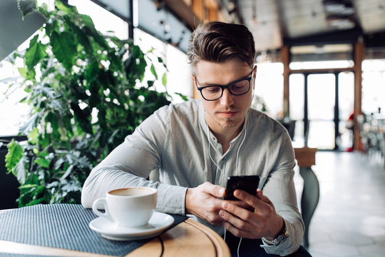 Pensive handsome man in headphones, watching or reading something on mobile phone, drinking coffee, having a break at cafe. Dressed in shirt.