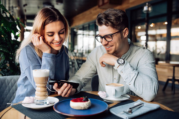 Young cheerful guy and girl in headphones, watching a movie on mobile phone while sitting at cafe and drinking coffee with dessert.