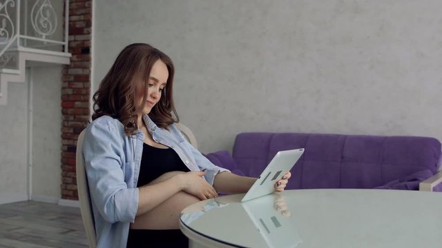 Happy pregnant woman sitting at a glass kitchen table drinking coffee and using a tablet computer. Engaged in business on the Internet. Remote work in the decree