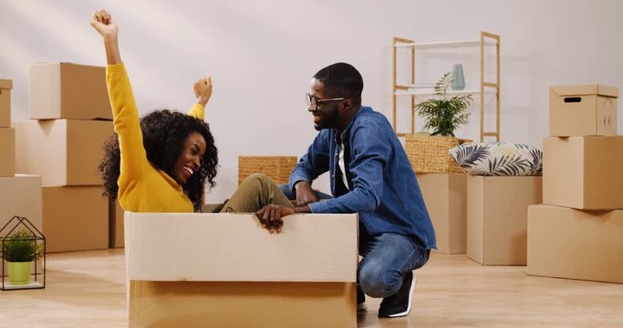 Cheerful African American couple moving in the new apartment. Curly woman sitting in the carton box and having fun with her girlfriend. Indoor