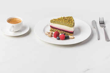 close up view of sweet cake with pistachios and berries on plate and cup of coffee isolated on white