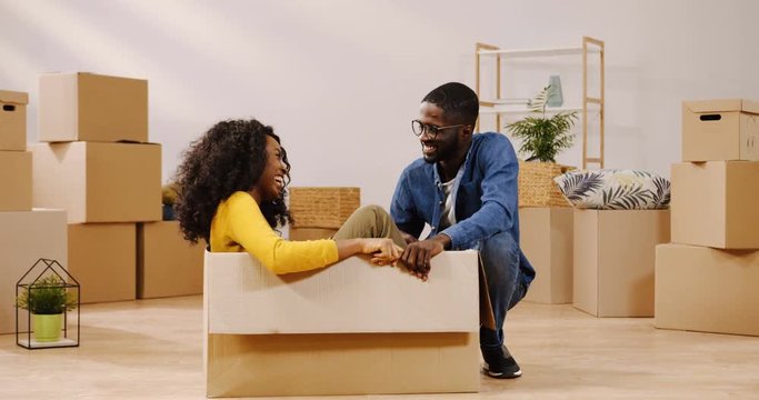 Young curly African American woman sitting in the carton box inthe middle of the cozy room among boxes, talking and laughing with her boyfriend. Indoors
