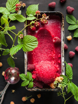 Raspberry sorbet (fruit ice cream scoop) in a container with raspberry berries and leaves on a black background with summer mood. Summer dessert top view. Bright summer refreshment.