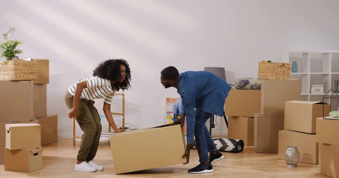 Young african american just married couple relocating a big carton box in the living room while moving in their home and woman feeling strong back pain. Inside