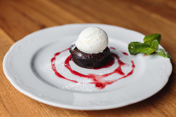 A horizontal photo of a small brownie cake with vanilla ice cream, decorated with mint, strawberry sauce and cinnamon, served on a white plate.