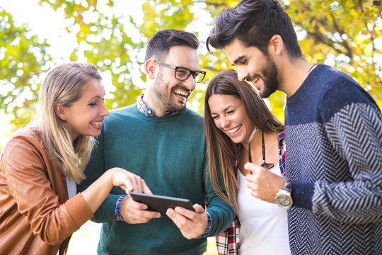 Image of four happy smiling young friends walking outdoors in the park holding digital tablet