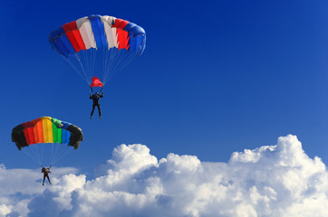 Fototapeta na wymiar two parachutists soar on colorful parachutes across the boundless blue sky against the background of white fluffy clouds