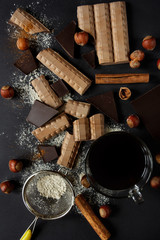 A cup of coffee on a black background. Pieces of milk and dark chocolate, hazelnuts, cinnamon and powdered sugar on a dark background. Flat lay.