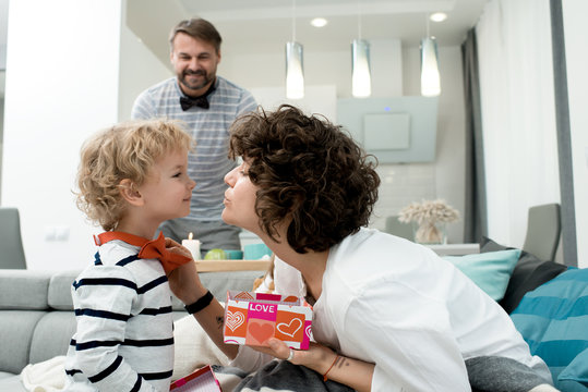 Side View Portrait Of Adorable Little Boy Giving Present To His Mom On Valentines Day, With Father In Background