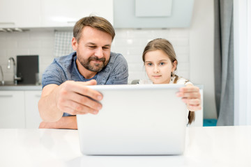 Portrait of smiling bearded man using laptop with daughter sitting at table in modern  kitchen