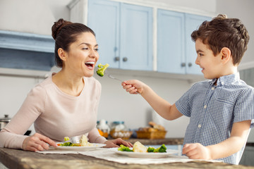 Happy together. Beautiful content dark-haired young mother smiling and eating healthy food with her son and her boy feeding her