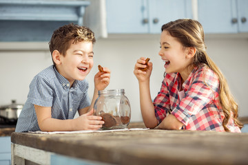 Joking. Cute happy little dark-haired brother and sister laughing and eating cookies while sitting at the table - obrazy, fototapety, plakaty