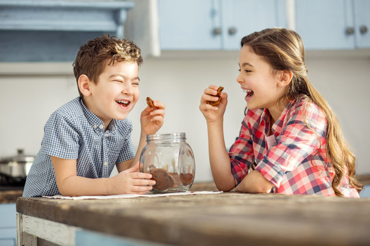 Joking. Cute happy little dark-haired brother and sister laughing and eating cookies while sitting at the table
