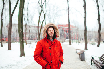 Portrait, close-up of a young stylishly dressed man smiling with a beard dressed in a red winter jacket with a hood and fur on his head. Winter and frost theme