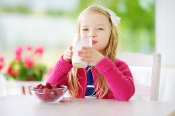 Pretty little girl eating raspberries and drinking milk at home. Cute child enjoying her healthy fresh organic fruits and berries.