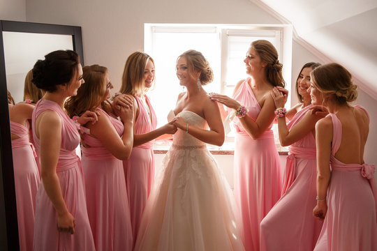 Bride In White Wedding Dress And Bridesmaids In Pink Dresses Posing In Hotel Or Fitting Room At Wedding Day.